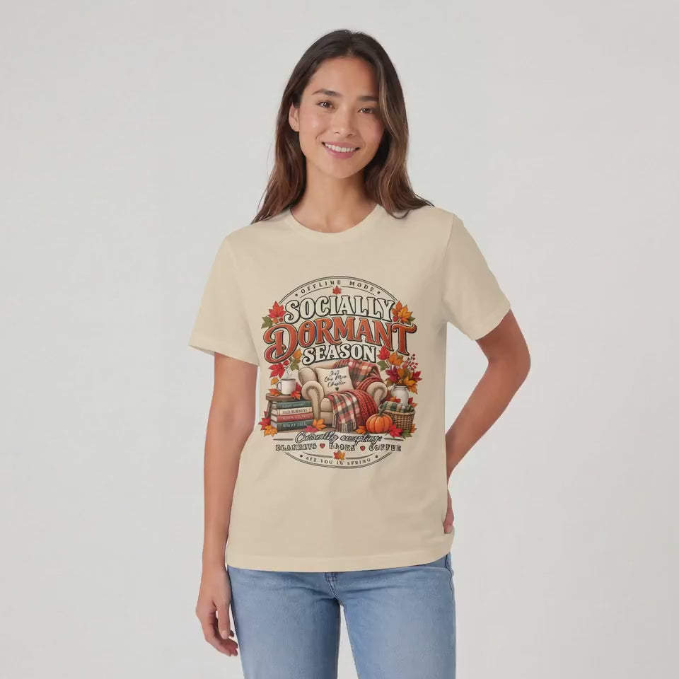 “Woman wearing a cream ‘Socially Dormant Season’ fall graphic T-shirt relaxes in a cozy autumn living room, reading a book by a fireplace with a warm plaid blanket, pumpkins, coffee, and stacks of books nearby.”